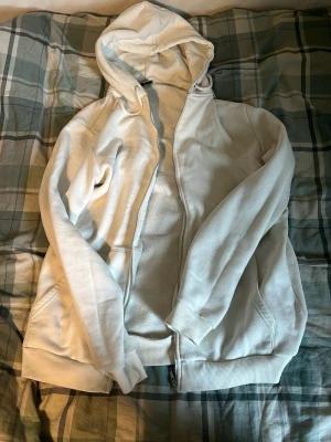 Vit zip hoodie med huva - Säljer en enkel och stilren vit hoodie med dragkedja och huva. Hoodien har fickor på sidorna och ribbade muddar vid ärmar och nederkant. Perfekt för chill dagar eller när du vill ha en clean look. Materialet känns mjukt och bekvämt.