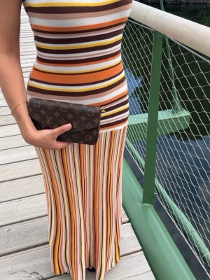 Randig långklänning  - Säljer en lång, ärmlös klänning med färgglada ränder i brunt, orange, gult, vitt och beige. Klänningen har en figurnära passform och är tillverkad i mjuk bomull, perfekt för varma dagar. Snygg och trendig design som sticker ut.
