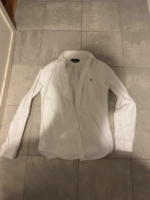 Vit skjorta från Ralph Lauren - Klassisk vit skjorta från Ralph Lauren med broderad logga på bröstet. Skjortan har lång ärm, knappar framtill och en tidlös design som passar till det mesta. Tillverkad i bomull för en skön känsla och snygg passform. Storlek S 