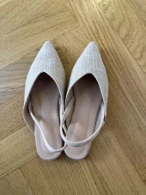 Beige ballerinaskor med spetsig tå - Säljer ett par stilrena beige ballerinaskor med spetsig tå och öppen häl. Skorna har en tunn rem runt hälen och är tillverkade i ett tygmaterial med diskret struktur. Perfekta för dig som gillar minimalistisk och trendig design.anvönda en gång endast.