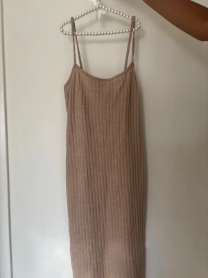 Beige ribbad långklänning barn strl 134 - Säljer en beige ribbad långklänning med smala axelband och rundad halsringning. Klänningen har en rak passform och är tillverkad i ett mjukt, stretchigt material som passar perfekt för varmare dagar. Med tillhörande tshirt