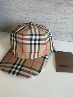 Beige rutig keps från Burberry - Säljer en klassisk beige keps från Burberry med det ikoniska rutmönstret i svart, vitt och rött. Kepsen har böjd skärm och justerbar rem bak. Tillverkad i ett mjukt och slitstarkt material som ger en lyxig känsla.