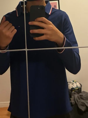 Blå långärmad skjorta Tommy Hilfiger S i EU - Snygg blå långärmad skjorta från Tommy Hilfiger med vita och röda detaljer på krage och ärmslut. Skjortan har klassisk krage och knappar framtill. Perfekt för dig som gillar stilrena plagg med en sportig touch.