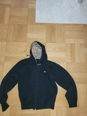 Svart hoodie med Burberry-mönster - Snygg svart hoodie med dragkedja och klassiskt Burberry-rutmönster i huvan. Har fickor framtill och liten broderad Burberry-logga på bröstet. Perfekt för dig som vill ha en stilren och exklusiv look.