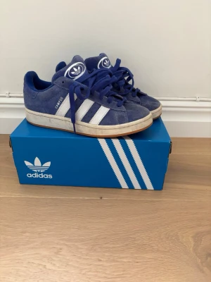 Adidas Campus blå sneakers - Säljer ett par klassiska Adidas Campus sneakers i blå mocka med vita detaljer och de ikoniska tre ränderna på sidan. Skorna har låga skaft och vit sula, samt Adidas-logga på plösen. Perfekta för dig som gillar retrostil och streetwear.