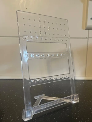 Transparent smyckesställ  - Snyggt och praktiskt smyckesställ i transparent akrylplast. Perfekt för att organisera och visa upp örhängen, ringar eller andra småsmycken. Stället har flera hål och två rader för enkel upphängning samt ett avtagbart stöd för stabilitet.