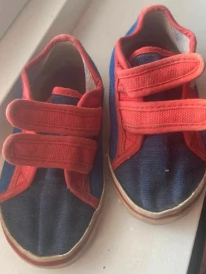 Blå och röda sneakers med kardborre (barn) - Säljer ett par blå sneakers med röda detaljer och två breda kardborreband. Skorna har vit sula och är gjorda i tygmaterial. Perfekt för dig som gillar färgstarka och bekväma skor med enkel stängning.
