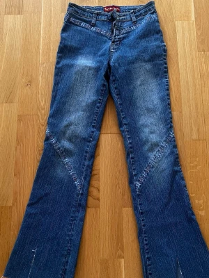 Blå bootcut jeans med brodyr - Säljer ett par blå bootcut jeans med snygg brodyrdetalj i blommigt mönster längs fickor och ben. Jeansen har klassisk femficksmodell, hällor för skärp och är tillverkade i denim. Perfekta för dig som gillar en retro och unik stil.