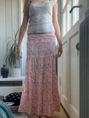 Långkjol med rosa paisleymönster - Säljer en flowy långkjol i vitt med rosa paisleymönster. Kjolen har bred resår i midjan och faller snyggt i flera lager. Perfekt för dig som gillar bohemisk stil och vill ha något bekvämt och luftigt.