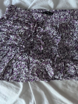 Lila blommig kortkjol med volang - Söt kortkjol med blommigt mönster i lila, vit och svart. Kjolen har volangdetaljer och resår i midjan för en bekväm passform. Perfekt för dig som gillar en feminin och trendig stil.