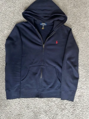 Mörkblå hoodie från Polo Ralph Lauren - Mörkblå hoodie med dragkedja från Polo Ralph Lauren i storlek 14-16. Klassisk design med röd broderad logga på bröstet, huva och känguruficka framtill. Perfekt för en chill och stilren look. 