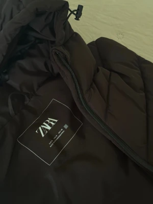 Svart dunväst med huva från ZARA - Snygg svart dunväst från ZARA med dragkedja och vadderad design. Storlek: S