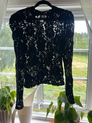 Svart långärmad spetsblus - Säljer en svart långärmad topp i genombruten spets med blommönster. Modellen har puffade axlar och rund halsringning. Perfekt för dig som gillar en edgy och elegant stil. Materialet är tunt och transparent, vilket ger en cool layering-effekt.