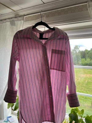 Rosa randig skjorta med knappar - Säljer en rosa skjorta med tunna vertikala blåa ränder och knappar framtill. Skjortan har lång ärm med bred manschett och en bröstficka. Perfekt för en avslappnad men ändå stilsäker look.