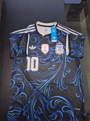Svart och blå Argentina fotbollströja - Säljer en svart och blå Argentina fotbollströja från Adidas med nummer 10, FIFA-märke och AFA-logga på bröstet. Tröjan har korta ärmar, vita detaljer och ett unikt blått mönster över hela plagget. Perfekt för dig som älskar fotboll och vill sticka ut på planen.