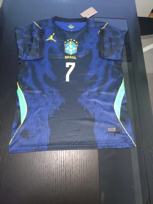 Blå Brasilien fotbollströja nr 7 - Snygg blå fotbollströja med Brasilien-emblem och nummer 7 på bröstet. Tröjan har gula och turkosa detaljer samt ett gult logotyptryck på höger sida. Tillverkad i ett lätt och ventilerande material, perfekt för träning eller match.