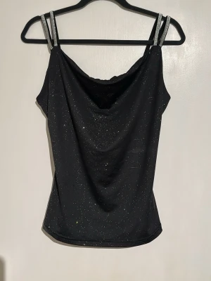Svart glittrig linnetopp med smala band - Snygg svart linnetopp med glittrigt tyg och dubbla smala axelband med silverdetaljer. Toppen har en draperad urringning och är figurnära i modellen. Perfekt för dig som vill sticka ut lite extra. Passar som Stl.  S oxå 