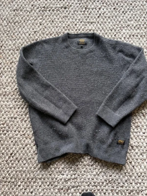 Mörkgrå stickad tröja Carhartt - Säljer en mörkgrå stickad tröja från Carhartt med rund halsringning och ribbade muddar vid ärmslut och nederkant. Tröjan har en diskret Carhartt-logga i gult och svart nertill. Perfekt för dig som gillar enkel och stilren design.