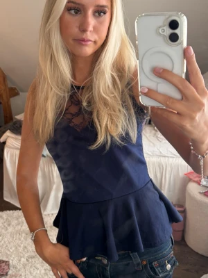 Spets topp - Snygg mörkblå ärmlös topp med volang nertill och transparent spets upptill. Toppen har rund halsringning och figurnära passform, perfekt för en trendig look.