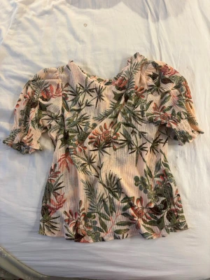 Blommig puffärm topp i ljusrosa - Supersöt topp med puffärmar i ljusrosa med blommigt mönster i grönt och rött. Ribbad struktur och kort ärm med volangkant. Perfekt för vår och sommar, ger en fräsch och trendig look till jeans eller kjol.