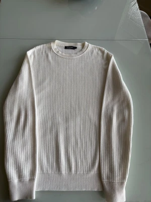 Vit stickad tröja med rund hals - Klassisk vit stickad tröja med rund hals och ribbade muddar vid ärmslut och nederkant. Tröjan har en enkel och stilren design med diskret struktur i stickningen. Perfekt för lager på lager och passar till det mesta.