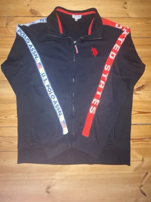 Svart zip-hoodie U.S. Polo Assn - Svart hoodie med dragkedja från U.S. Polo Assn. Röd logga på bröstet och text längs ärmarna, ena sidan med röd 'United States' och andra med blå 'U.S. Polo Assn'. Snygga detaljer och fickor med dragkedja. Perfekt för en sportig och avslappnad stil.
