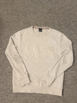 Ljusgrå sweatshirt från Hugo Boss - Snygg ljusgrå sweatshirt från Hugo Boss med diskret broderad logga framtill. Tröjan har rund halsringning, långa ärmar och ribbade muddar vid ärmslut och nederkant. Perfekt för en clean och stilren look.