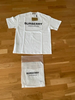 Vit Burberry t-shirt med logga - Säljer en vit t-shirt från Burberry med stor svart logga och texten 'London England' på bröstet. Klassisk rund halsringning och korta ärmar. Tillverkad i mjuk bomull och levereras med originalpåse och tagg. Står storlek S men är M