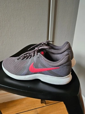 Nike  - Snygga grå Nike med en tydlig rosa märke på sidan. Skorna har en vit, tjock sula och är gjorda i meshmaterial. Klassisk låg modell med snörning, perfekt för träning eller chill vardagsstil.