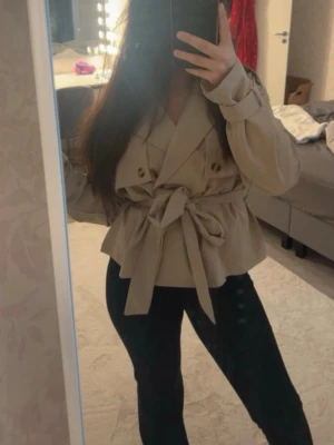Beige trenchcoat med knytskärp - Stilren beige trenchcoat med dubbelknäppning och brett knytskärp i midjan. Jackan har klassisk krage, dekorativa axelklaffar och vida ärmar. Perfekt för dig som vill ha en trendig och tidlös look till hösten.