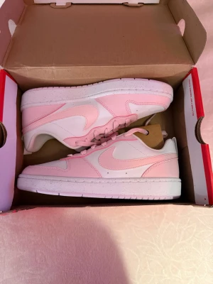 Nike rosa och vita sneakers - Säljer ett par snygga Nike sneakers i lågt modell med ljusrosa och vita paneler. Skorna har klassisk swoosh på sidan, perforerad tå och vita sulor. Tillverkade i läder och textil för en fräsch look. Perfekta för dig som gillar stilrena och trendiga sneakers.