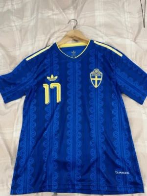 Blå Sverige fotbollströja nr 17 - Säljer en blå fotbollströja från Adidas med Sveriges landslagsemblem och nummer 17 i gult. Tröjan har GYÖKERES tryckt på ryggen, gula detaljer på axlarna och CLIMACOOL-material för bra ventilation. Perfekt för match eller träning.