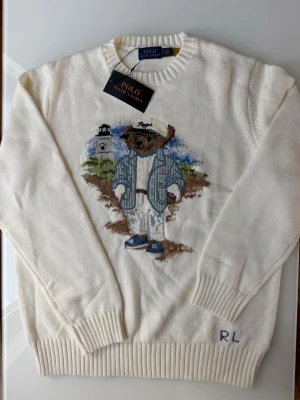 Vit stickad tröja Polo Ralph Lauren - Vit stickad tröja från Polo Ralph Lauren med ikonisk björn i blå jacka och keps broderad på framsidan. Tröjan har ribbade muddar och rund hals. Materialet är mjukt och passar perfekt för en avslappnad men stilren look.