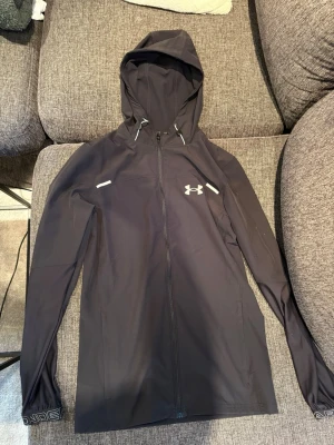 Svart vindjacka från Under Armour - Snygg svart vindjacka från Under Armour med huva och dragkedja framtill. Jackan har logga på bröstet, justerbar huva med snören och diskreta reflexdetaljer. Perfekt för blåsiga dagar och sportiga outfits.