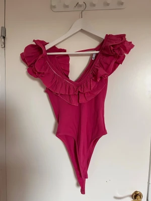 Rosa body med volangdetaljer - Snygg rosa body med djup urringning och stora volanger runt axlar och halsringning. Tillverkad i mjukt stretchigt tyg som sitter bekvämt mot kroppen. Perfekt för dig som vill ha något extra till din outfit.
