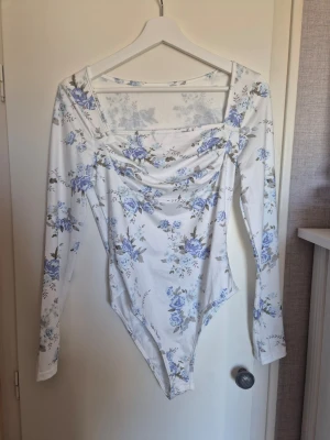 Vit body med blå blommor och långa ärmar - Säljer en vit body med blått blommönster och långa ärmar. Bodyn har en snygg drapering framtill och långa ärmar. Materialet ser ut att vara stretchigt och mjukt, perfekt för en trendig look. Passar dig som gillar romantiska detaljer och blommiga mönster. Aldrig använd