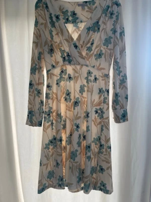 Blommig omlottklänning med långa ärmar - Söt kortklänning med blommigt mönster i grönt och beige på vit botten. Klänningen har omlott-skärning, v-ringning och långa ärmar. Perfekt för vår och sommar, med en lätt och luftig känsla.