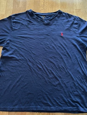 Marinblå t-shirt från Polo Ralph Lauren - Snygg marinblå t-shirt från Polo Ralph Lauren med klassisk v-ringning och röd broderad logga på bröstet. Tillverkad i mjuk bomull och har en normal passform. Perfekt för en clean och stilren look.