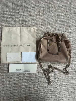 Beige Stella McCartney väska - Säljer en beige Stella McCartney handväska med kedjor som var guldiga innan men väldigt slitna och har tappat färg. Det medkommer dustbag och kort på bild. Den har ett hål i innerfodret och en vattenfläck på utsidan. Den är ganska sliten och i begagnat skick. Köptes på Plick. 