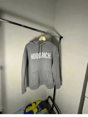 Grå HOODRICH hoodie med tryck - Snygg grå hoodie med stor vit HOODRICH-logga på bröstet. Hoodien har huva med dragsko och en stor magficka framtill. Tillverkad i mjukt material som känns skönt mot huden. Perfekt för en avslappnad och trendig stil.