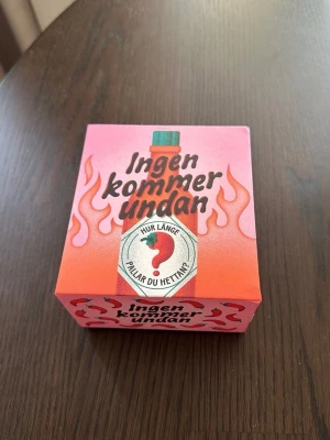 Sällskapsspel Ingen kommer undan - Roligt sällskapsspel i en färgglad rosa och orange låda med eldflammor och chili-motiv. Spelet heter 'Ingen kommer undan' och handlar om att testa hur länge man klarar av hetta. Perfekt för spelkvällar med vänner som gillar utmaningar och skratt.