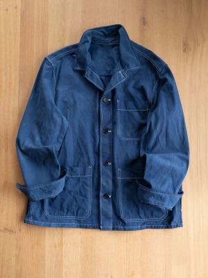 Marinblå Chore Jacket med knappar - Snygg marinblå overshirt i bomull med kontrasterande vita sömmar och tre stora fickor framtill. Jackan har klassisk krage, knäppning med svarta knappar och uppvikta ärmar för en avslappnad look.