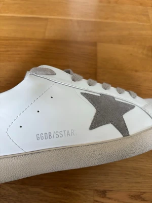 Golden Goose Super-Star vita sneakers - Golden Goose Super-Star sneakers i vitt läder med grå mockadetaljer och ikonisk stjärna på sidan. Skorna har grå snören och en klassisk låg siluett. Snygg GGDB/SSTAR-tryck på sidan och bekväm innersula. Perfekta för en trendig och avslappnad stil.