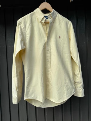Gul skjorta från Ralph Lauren - Klassisk ljusgul skjorta från Ralph Lauren med button-down krage och broderad logga på bröstet. Skjortan har lång ärm, normal passform och är tillverkad i mjuk bomull. Perfekt för en clean och stilren look.