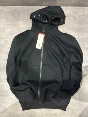 Svart hoodie med glasögon från C.P. Company - Säljer en svart hoodie från C.P. Company med hel dragkedja och ikoniska glasögon i huvan. Hoodien har ribbade muddar, två fickor framtill och diskret logga på bröstet. Tillverkad i mjukt bomullsmaterial för en skön och avslappnad look.