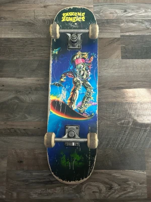 Skateboard Extreme Zombies motiv - Cool skateboard med färgstarkt Extreme Zombies-motiv på undersidan. Brädan har en blå bakgrund med en zombiefigur i neonfärger och texten 'Extreme Zombies' i gult. Utrustad med vita hjul och metalltruckar. Perfekt för dig som gillar unika brädor och streetstil.