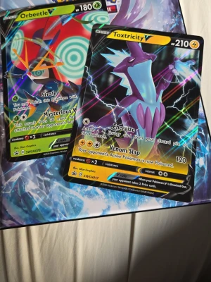 Samling Pokémonkort i pärm och lösa - Säljer en samling Pokémonkort med flera holografiska och VMAX-kort, bland annat Charizard, Flareon, Pikachu och Solgaleo. Korten har färgstarka illustrationer och vissa är regnbågsglittriga. Perfekt för dig som vill utöka din samling eller börja samla Pokémonkort. Lägg gärna bud, räknat ihop allt och sammanlagt ligger det runt 3700-ca4000. Pärmen ingår!!! Finns även en till likadan pärm som man kan få med för lite extra. Sleeves ingår!