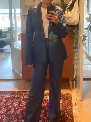 Mörkblå kostym från NA-KD - Snygg mörkblå glittrig kostym från NA-KD med klassisk kavaj och matchande byxor. Kavajen har två knappar och inte riktiga fickor i storlek 36. Byxorna är raka och har hög midja och riktiga fickor i storlek 32. OBSERVERA att byxorna är i mindre storlek än kavajen! Jag är 175 cm för referens! 