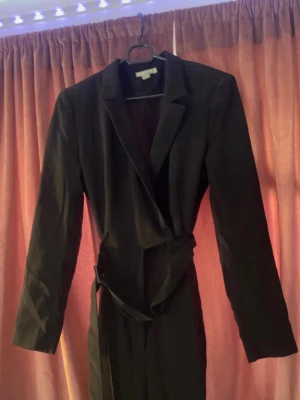 Svart långärmad jumpsuit med skärp - Stilren svart jumpsuit med långa ärmar och klassisk krage. Modellen har ett knytbart tygskärp i midjan som ger en snygg siluett. Passar perfekt till dig som gillar en clean och elegant look. Materialet ser ut att vara mjukt och följsamt.