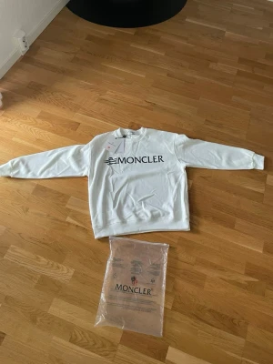 Vit sweatshirt från Moncler - Säljer en stilren vit sweatshirt från Moncler med stor svart logga på bröstet. Tröjan har rund halsringning, långa ärmar och ribbade muddar vid ärmslut och nederkant. Perfekt för dig som gillar exklusiva streetwear-plagg.
